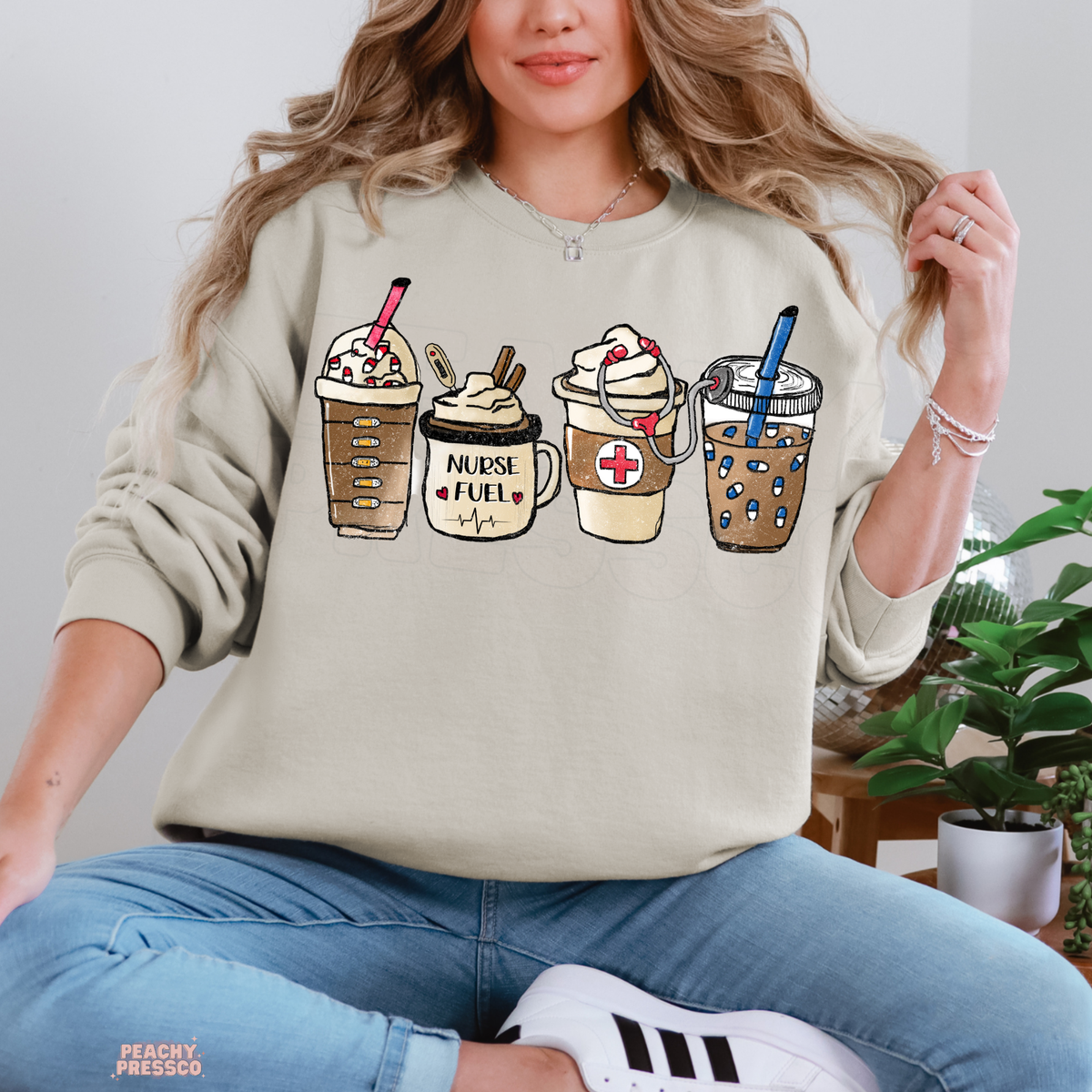 Sand Nurse Fuel coffee sweatshirt featuring iced coffee, a Nurse Fuel mug, a medical cross and stethoscope, paired with blue jeans.
