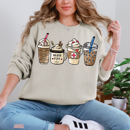 Sand Nurse Fuel coffee sweatshirt featuring iced coffee, a Nurse Fuel mug, a medical cross and stethoscope, paired with blue jeans.