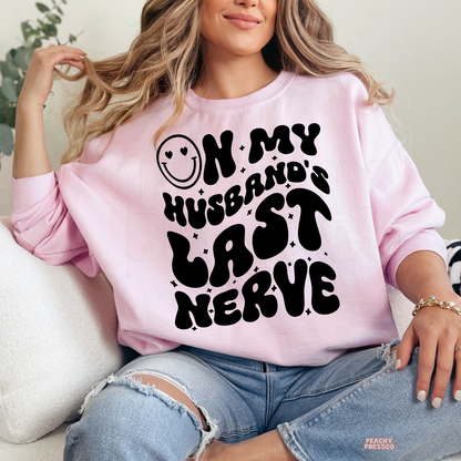 Light pink sweatshirt featuring a humorous "On My Husband's Last Nerve" graphic with oversized retro lettering. Cute wife gift for anniversaries or birthdays.