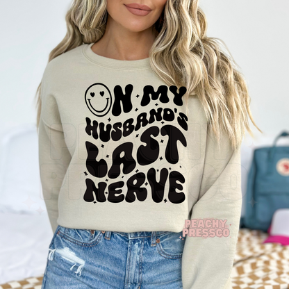 Sand sweatshirt featuring a bold "On My Husband's Last Nerve" graphic with retro bubble lettering and smiley face. Funny wife sweatshirt for everyday wear.