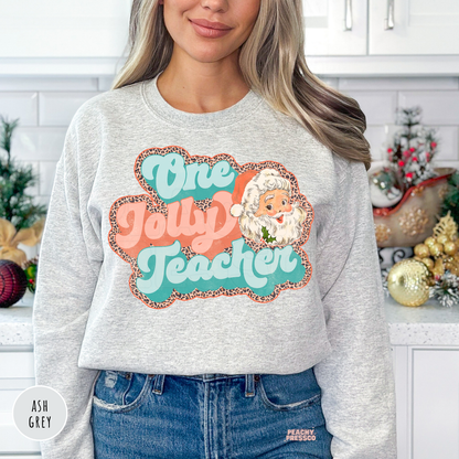 A woman wearing a ash grey sweatshirt with a colorful graphic that reads 'One Jolly Teacher' in teal and coral with a leopard-print border and a Santa face. 