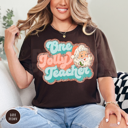 A woman wearing a dark brown graphic t-shirt with a colorful 'One Jolly Teacher' design featuring a cartoon Santa Claus, paired with light blue jeans.