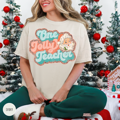A woman wearing an ivory graphic t-shirt that reads 'One Jolly Teacher' in teal and coral with a cartoon Santa illustration, paired with green pants, sitting in front of decorated Christmas trees.