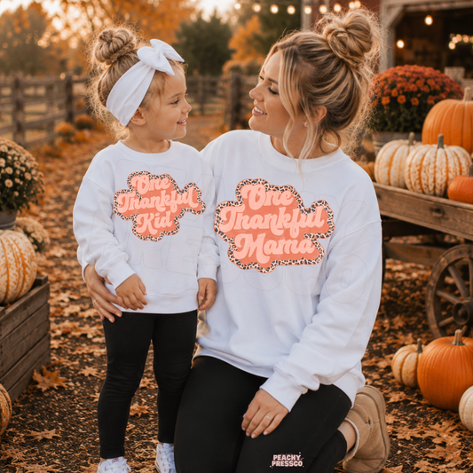 Mom and daughter wearing matching white One Thankful Mama and One Thankful Kid sweatshirts at a pumpkin patch during a cozy fall day.