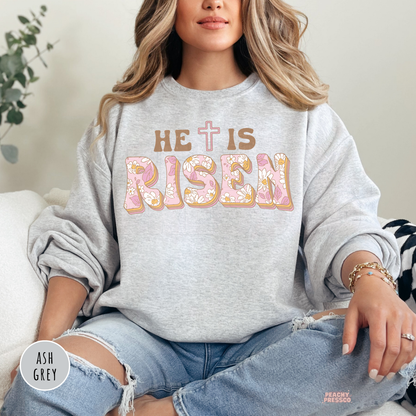 Ash grey crewneck sweatshirt featuring a vintage floral "He Is Risen" design with pastel flower-filled lettering and a cross. Faith-inspired Christian sweatshirt for Easter and spring.