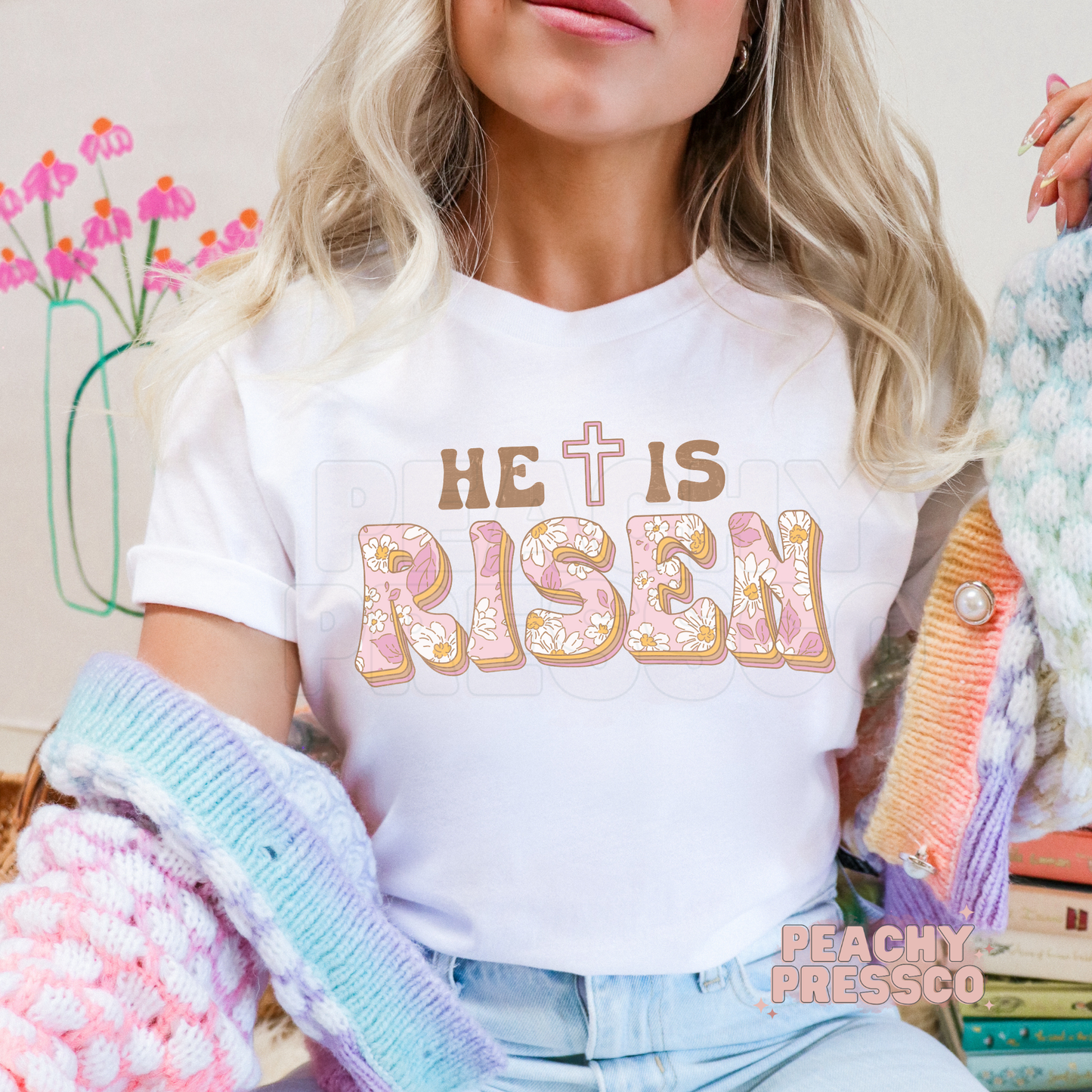 White unisex t-shirt with a retro "He Is Risen" floral graphic featuring pastel pink floral letters, brown text, and a cross. Cute Christian Easter shirt for women and everyday faith wear.