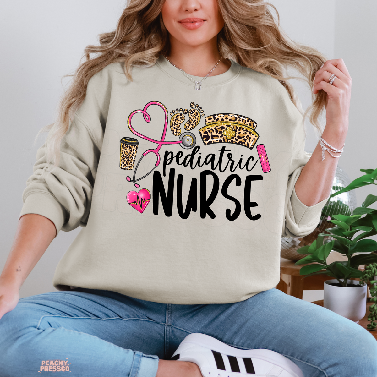 Sand pediatric nurse crewneck sweatshirt featuring a pink heart-shaped stethoscope, leopard baby footprints, nurse hat and pink heartbeat heart.