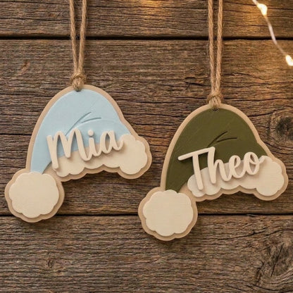 Light blue and green personalized Santa hat Christmas ornaments hanging on a rustic wooden background with the names Mia and Theo