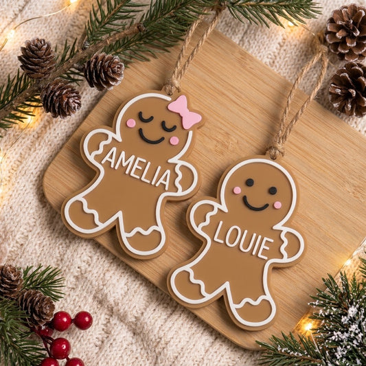 Two personalized gingerbread man Christmas ornaments on a wooden board, surrounded by pine branches, pinecones, and warm fairy lights. One ornament is with a pink bow, and the other is plain.