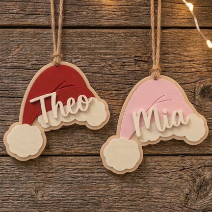 Red and pink personalized Santa hat Christmas ornaments hanging on a rustic wooden background with the names Theo and Mia