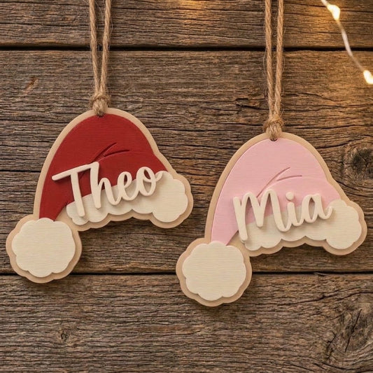 Red and pink personalized Santa hat Christmas ornaments hanging on a rustic wooden background with the names Theo and Mia