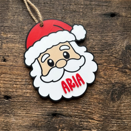 A custom personalized Santa Claus face Christmas ornament with the name Aria on the fluffy white beard, hanging on a rustic wooden background