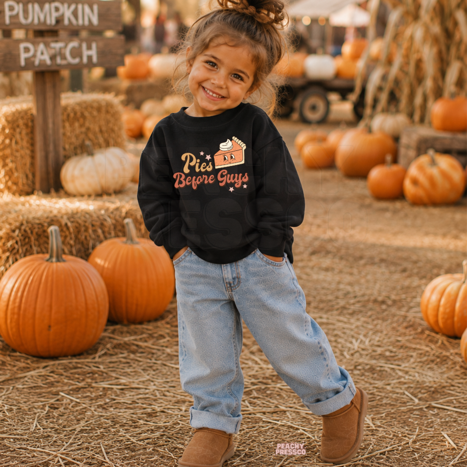 Happy toddler girl wearing a black "Pies Before Guys" sweatshirt at a pumpkin patch surrounded by pumpkins and warm autumn decorations.