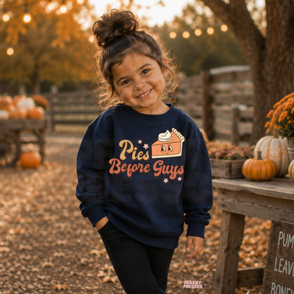 Toddler girl wearing a navy "Pies Before Guys" sweatshirt at a pumpkin patch with pumpkins, autumn leaves, and rustic fall decorations.