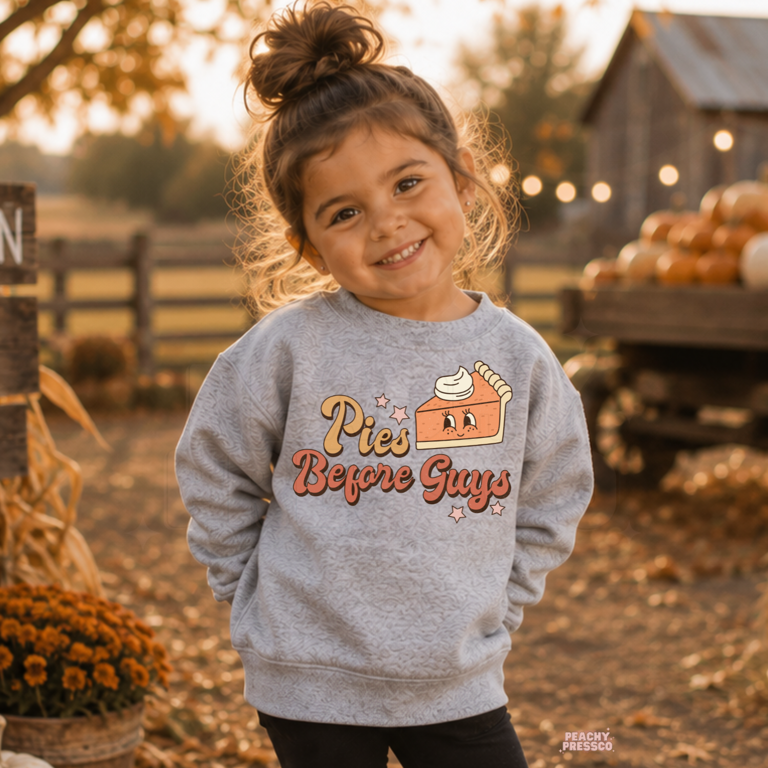 Toddler girl smiling while wearing a sport grey "Pies Before Guys" sweatshirt at a pumpkin patch with pumpkins and cozy fall décor.