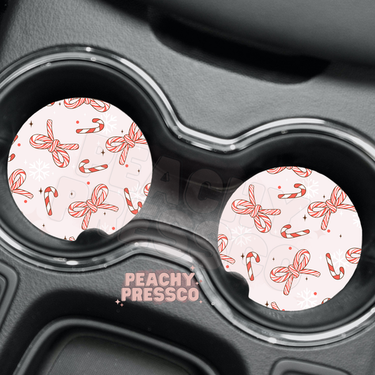 Pair of light pink car coasters with red striped candy cane ribbon bows and snowflake pattern inside car cup holders.