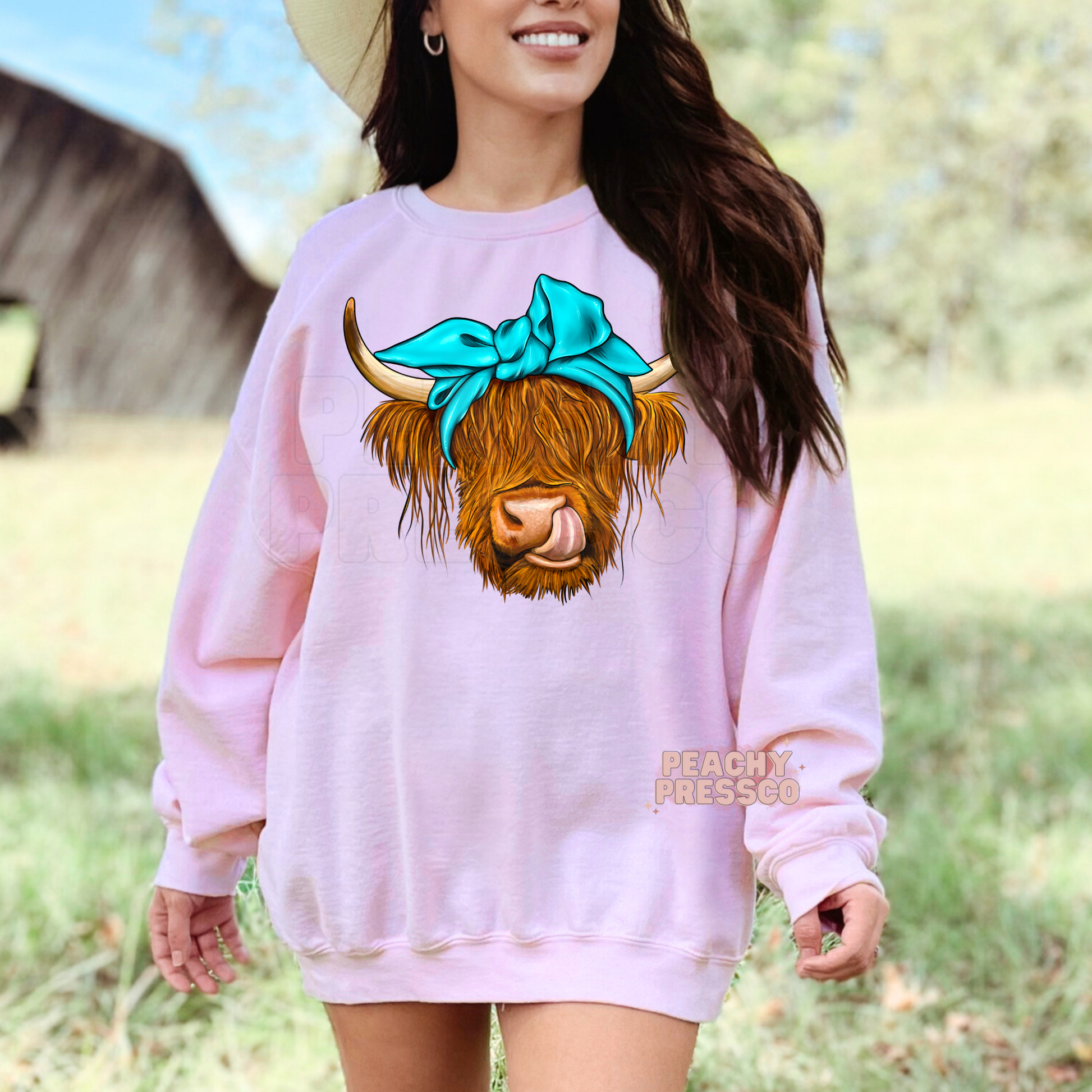 Woman wearing a pink sweatshirt with a cute Highland cow wearing a turquoise bow and sticking its tongue out, perfect for country girls.