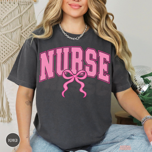 Pepper charcoal nurse graphic T-shirt with large pink collegiate NURSE text and a cute ribbon bow, styled with blue jeans.
