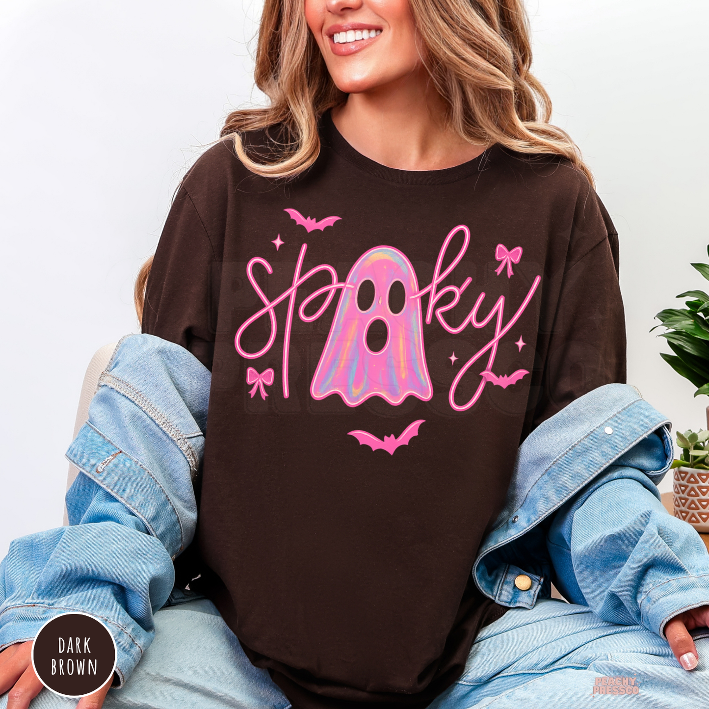 Woman wearing a dark brown crewneck sweatshirt featuring a trendy pink spooky ghost Halloween design with bows and bats.