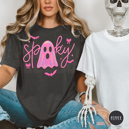 Woman wearing a Comfort Colors Pepper t-shirt with a trendy pink spooky ghost design beside a Halloween skeleton decoration.