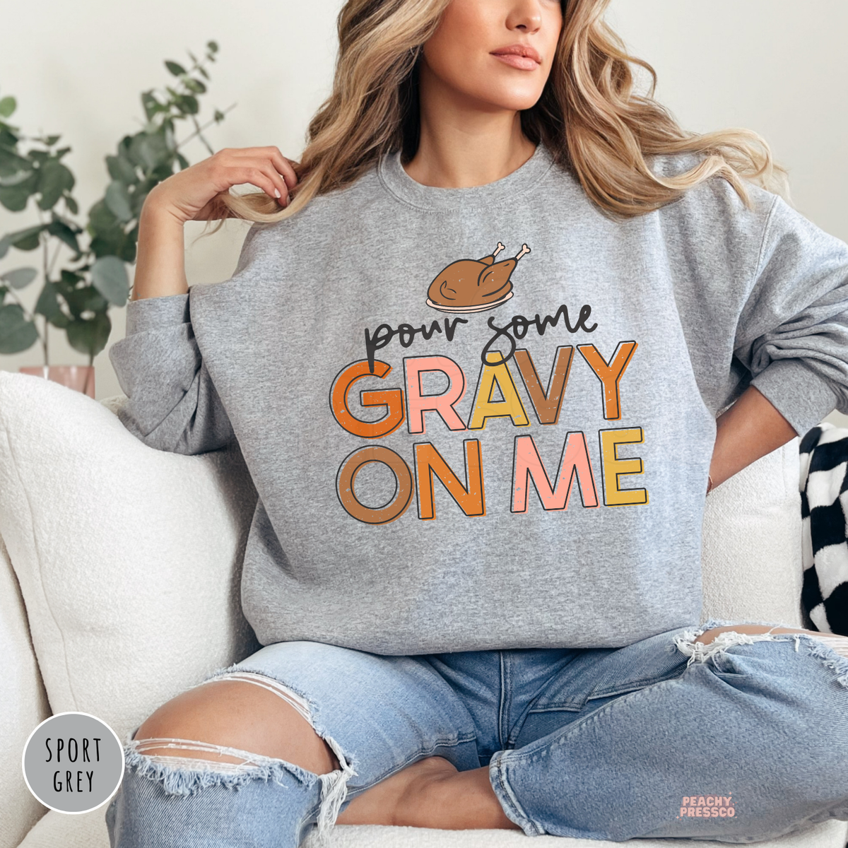 Woman wearing a sport grey crewneck sweatshirt featuring a funny "Pour Some Gravy On Me" Thanksgiving graphic with colorful retro lettering and a roast turkey.