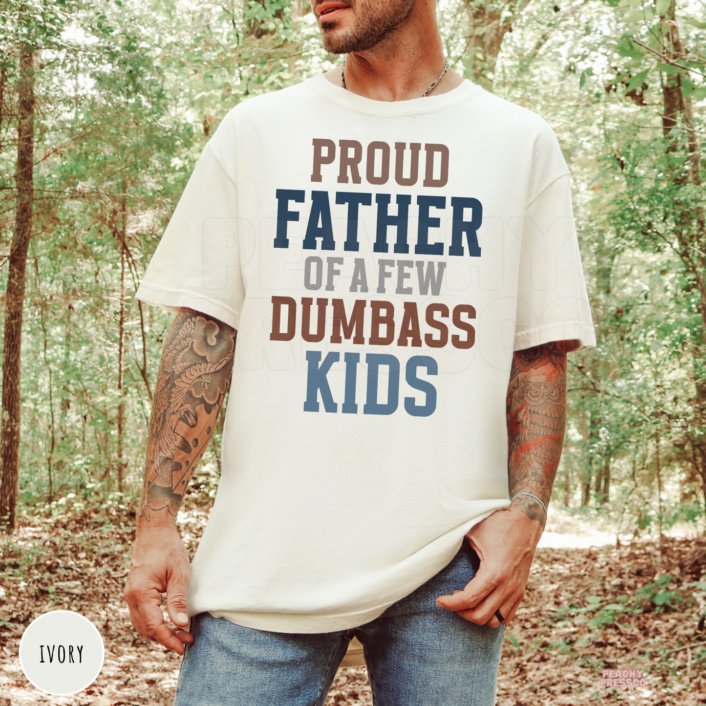 Man wearing an ivory Comfort Colors t-shirt featuring the "Proud Father of a Few Dumbass Kids" graphic outdoors in a wooded setting.