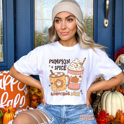 Woman wearing a white graphic t-shirt with a playful "Pumpkin Spice & Everything Nice" design featuring a pumpkin spice coffee, hot cocoa, and cute autumn drink illustrations.