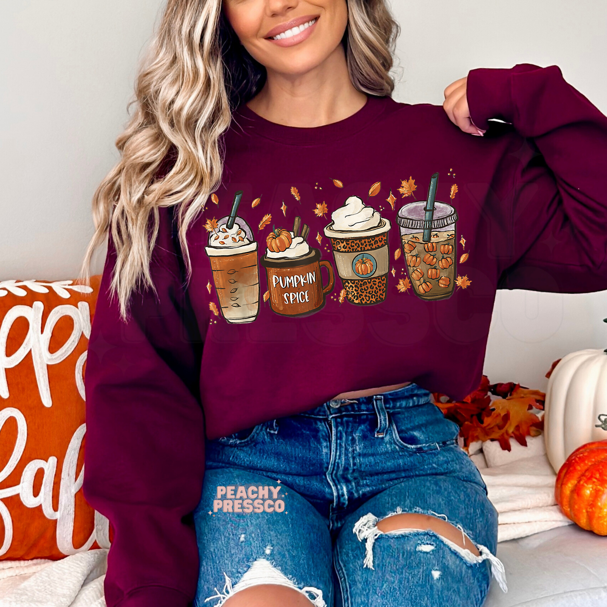 Woman wearing a maroon crewneck sweatshirt featuring a cute pumpkin spice latte coffee graphic with cozy fall drinks and autumn leaves.
