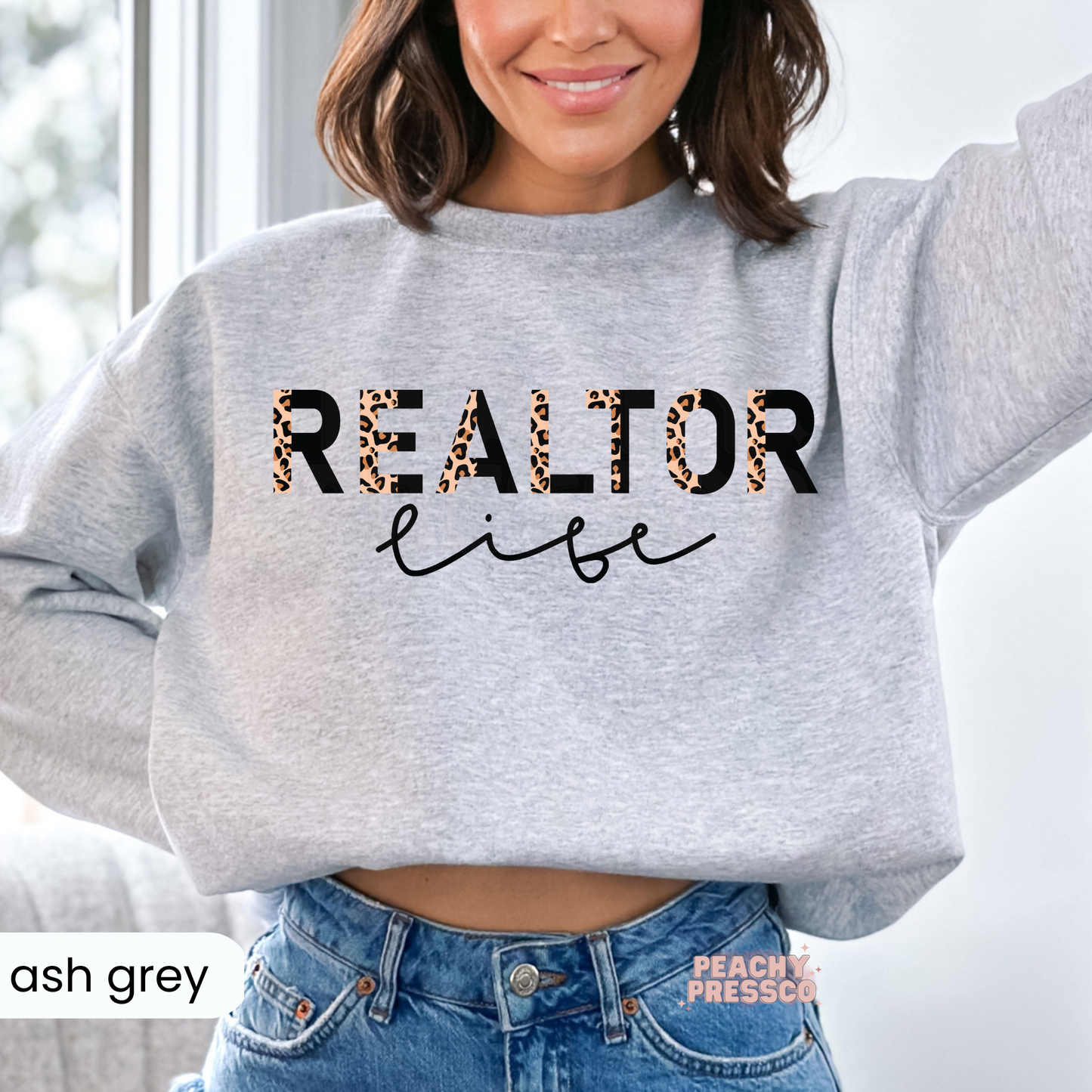 Woman wearing an ash grey sweatshirt with a Realtor Life leopard print graphic, cozy apparel for REALTORS®, brokers, and real estate agents.