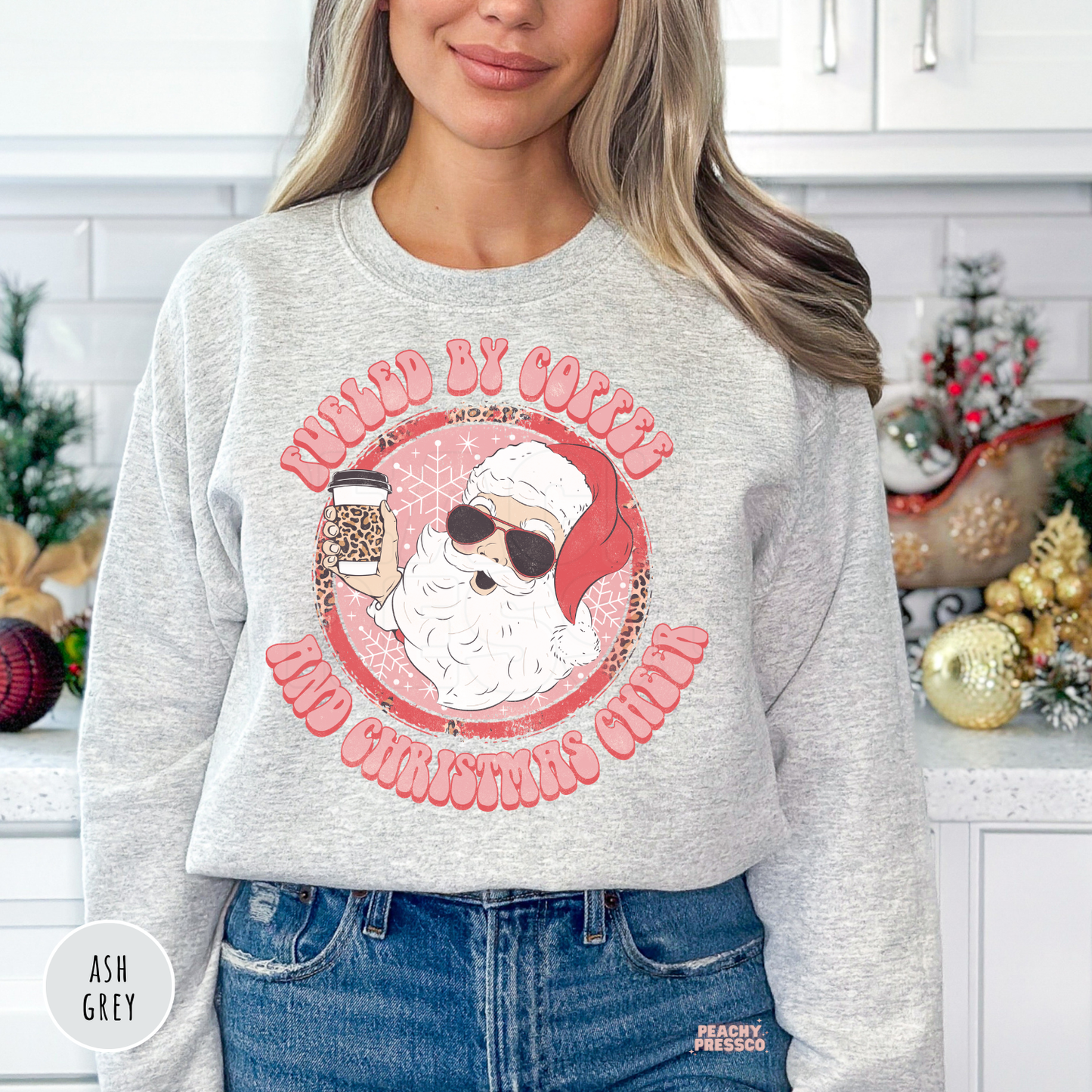 Woman standing in a kitchen wearing an ash grey crewneck sweatshirt featuring a pink retro Santa wearing sunglasses with Fueled By Coffee And Christmas Cheer graphic.