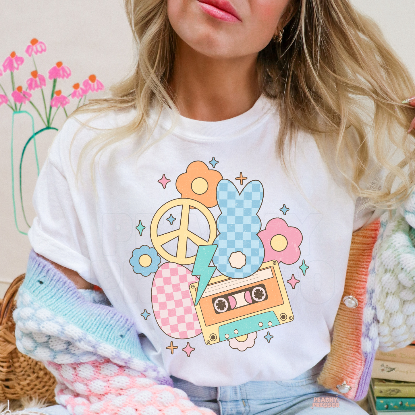 Woman wearing a white t-shirt featuring a retro pastel Easter graphic with a bunny, cassette tape, peace sign, flowers, and lightning bolt.