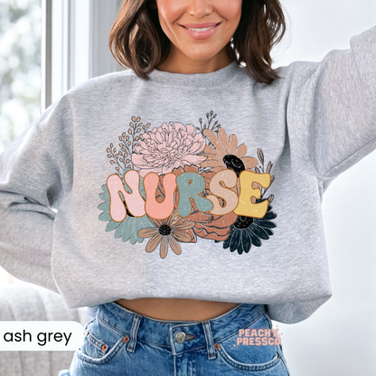 Woman wearing an ash grey retro floral nurse sweatshirt with vintage pastel floral artwork and colorful "NURSE" text.