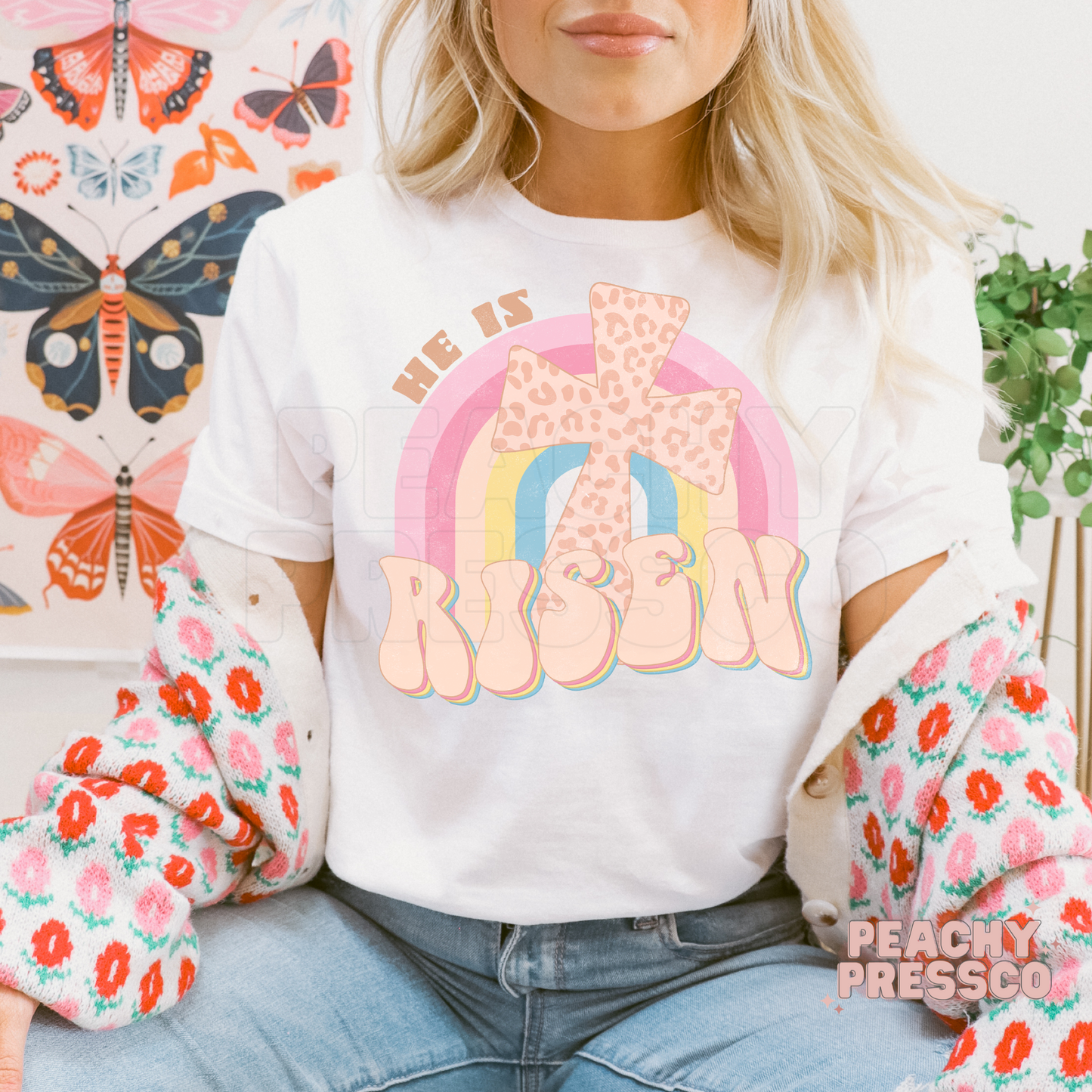 White unisex t-shirt featuring a retro "He Is Risen" design with a leopard-print cross, pastel rainbow, and vintage bubble lettering. Christian Easter shirt celebrating the resurrection of Jesus.