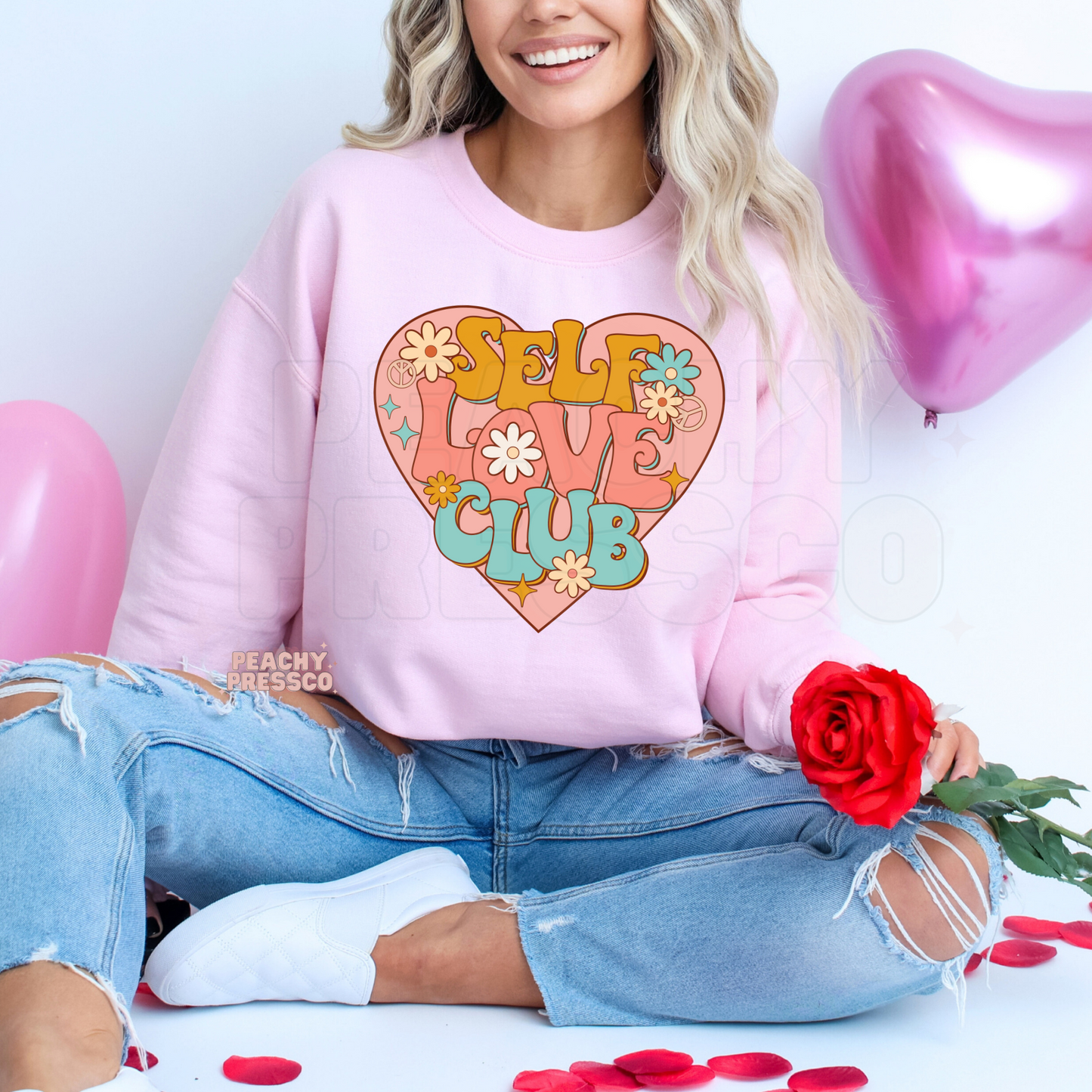 Light pink sweatshirt featuring a colorful Self Love Club heart graphic with groovy lettering, floral details, and a retro aesthetic.