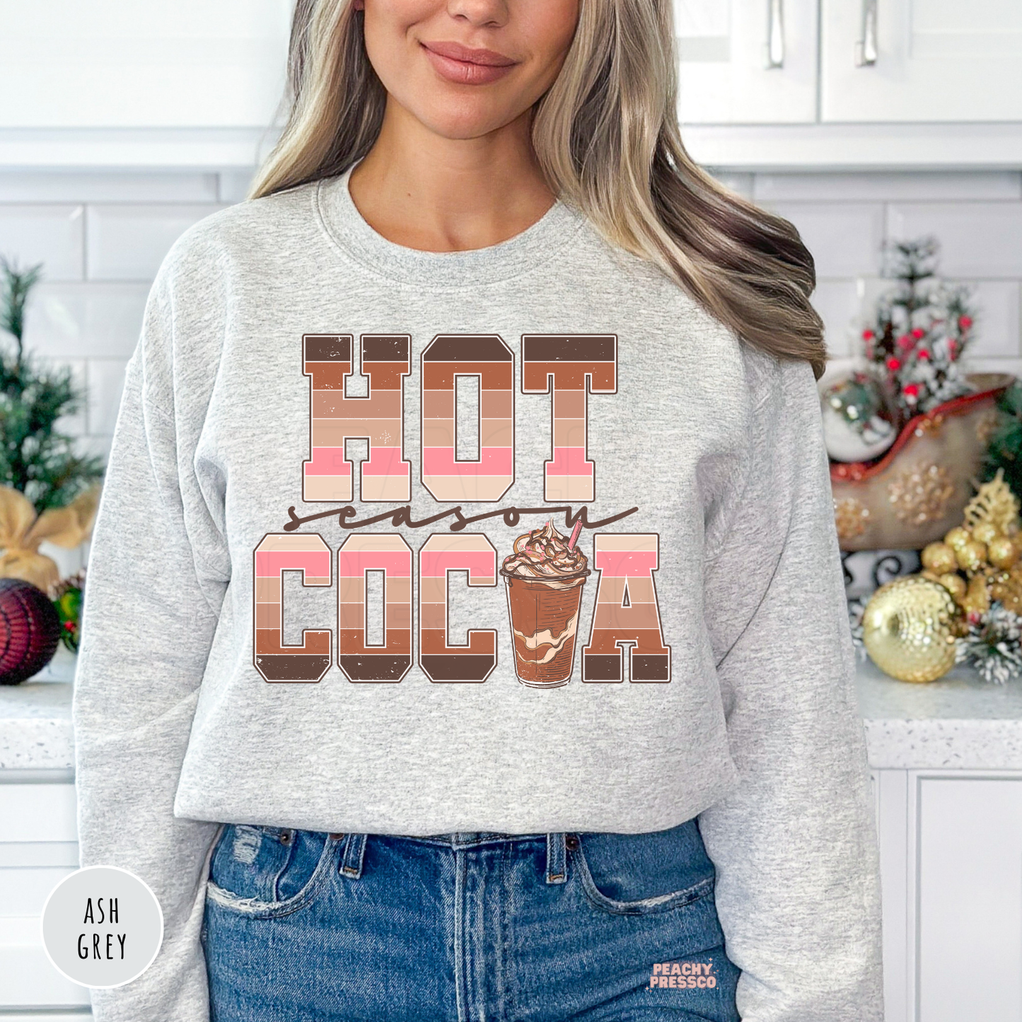 Woman wearing an ash grey Hot Cocoa Season sweatshirt featuring retro striped lettering and a hot chocolate cup graphic.