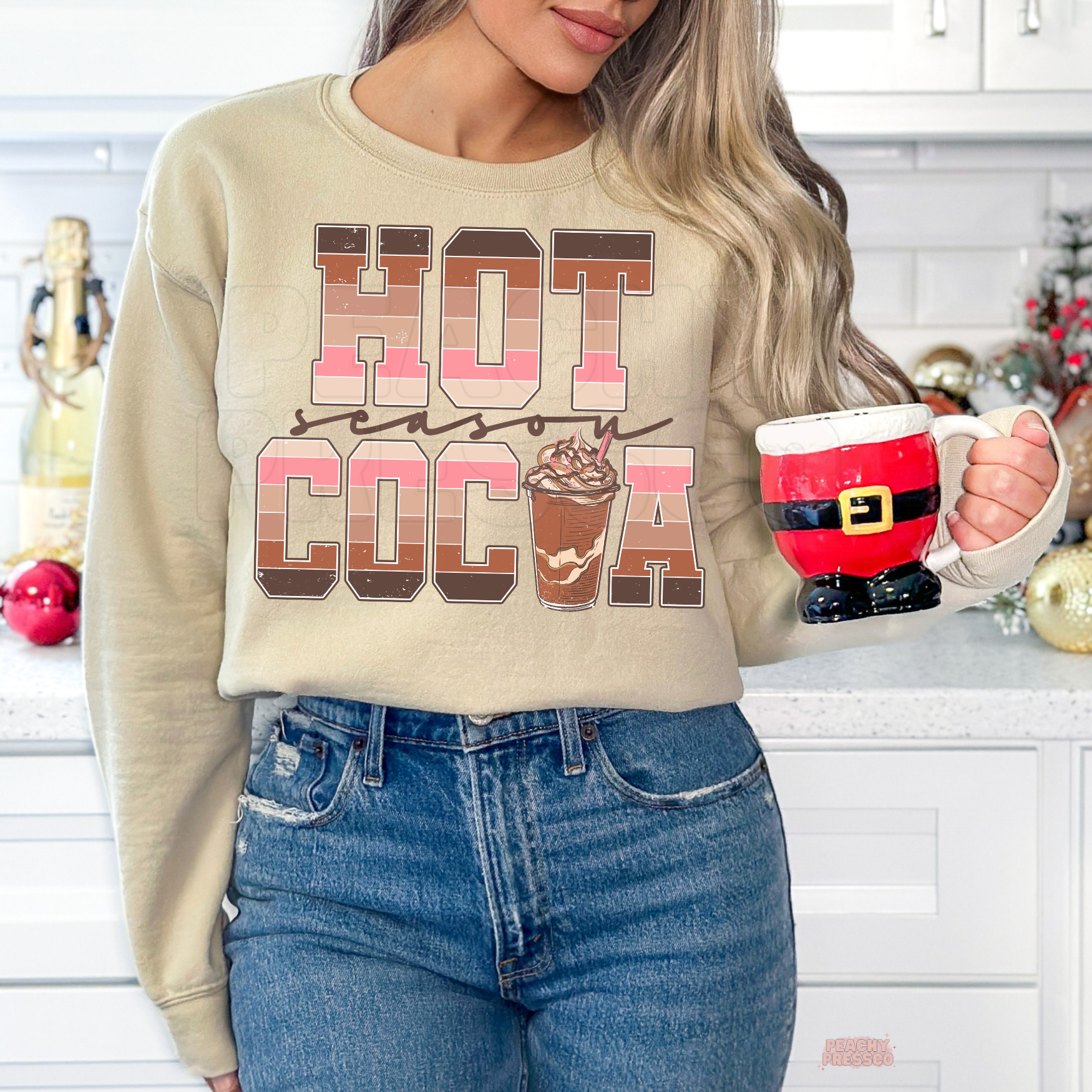 Woman wearing a sand beige Hot Cocoa Season crewneck sweatshirt holding a red Santa Claus mug in a bright kitchen.