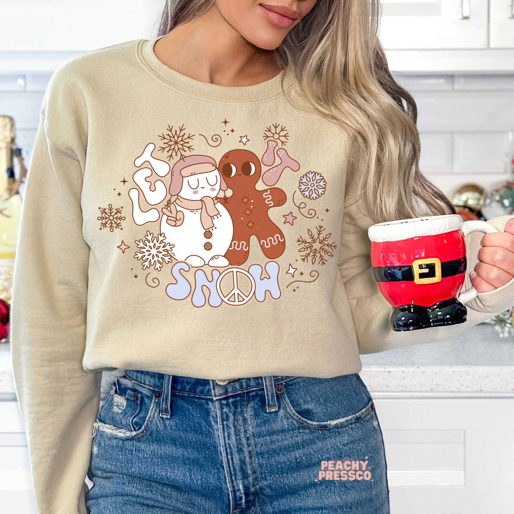 Woman wearing a sand Let It Snow sweatshirt holding a Santa Claus boot mug in a festive kitchen.