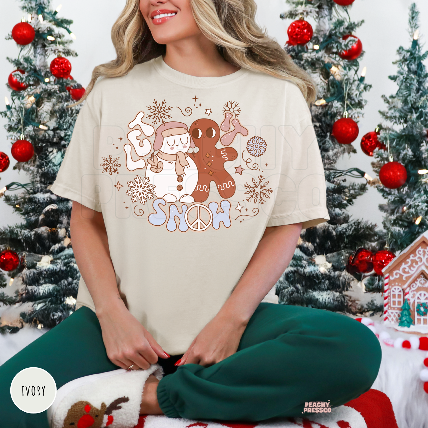 Woman wearing an ivory retro Let It Snow t-shirt featuring a cute snowman and gingerbread man graphic sitting in front of Christmas trees.