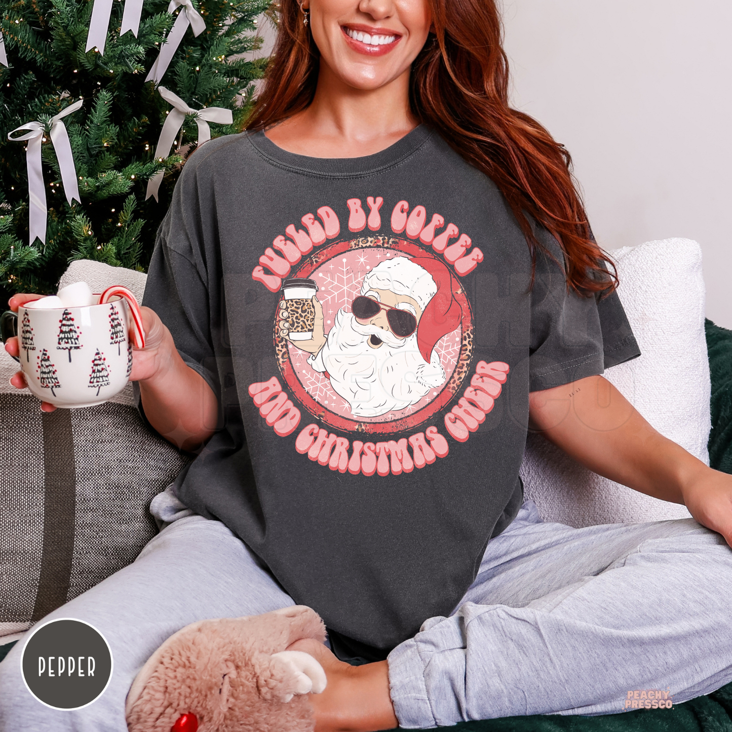 Woman holding a mug with marshmallow and candy cane sitting on a couch wearing a pepper grey t-shirt with Fueled By Coffee And Christmas Cheer design.