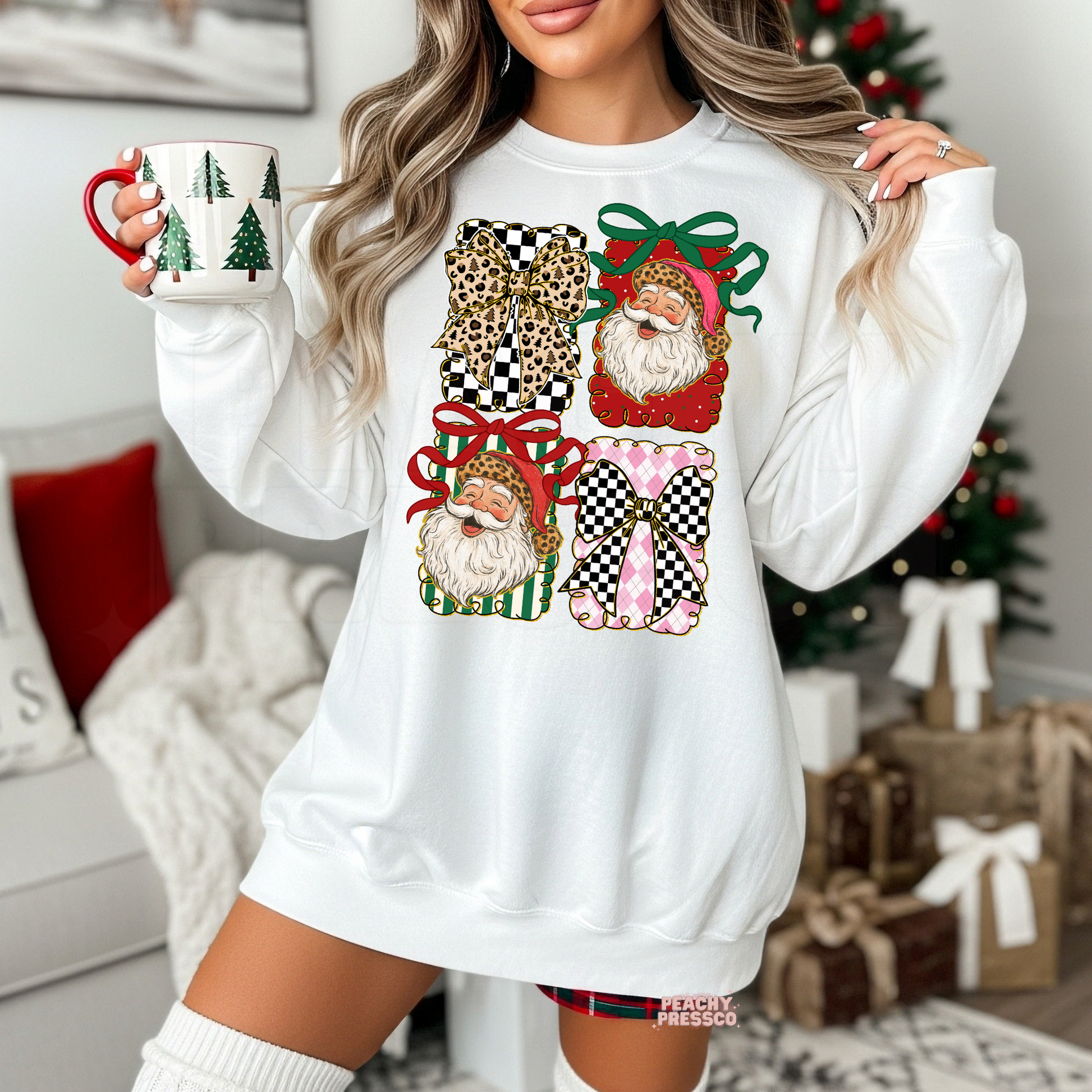 A woman wearing a white oversized sweatshirt with a festive Christmas graphic print featuring Santa in gift boxes. She is holding a mug in a cozy living room setting with a Christmas tree and wrapped presents in the background.