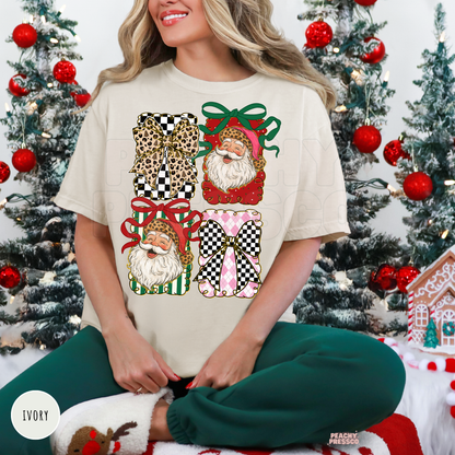 A woman wearing an ivory graphic t-shirt with a Christmas-themed gift box print, sitting in front of decorated Christmas trees. The t-shirt features a graphic of four gift boxes with leopard print, checkered patterns, and Santa Claus faces with red and green bows.