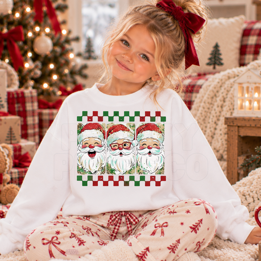 A young girl with blonde hair in a red bow sits cross-legged in a cozy Christmas living room, wearing a white crewneck sweatshirt with a graphic of three Santa Claus faces and matching patterned pajama pants.