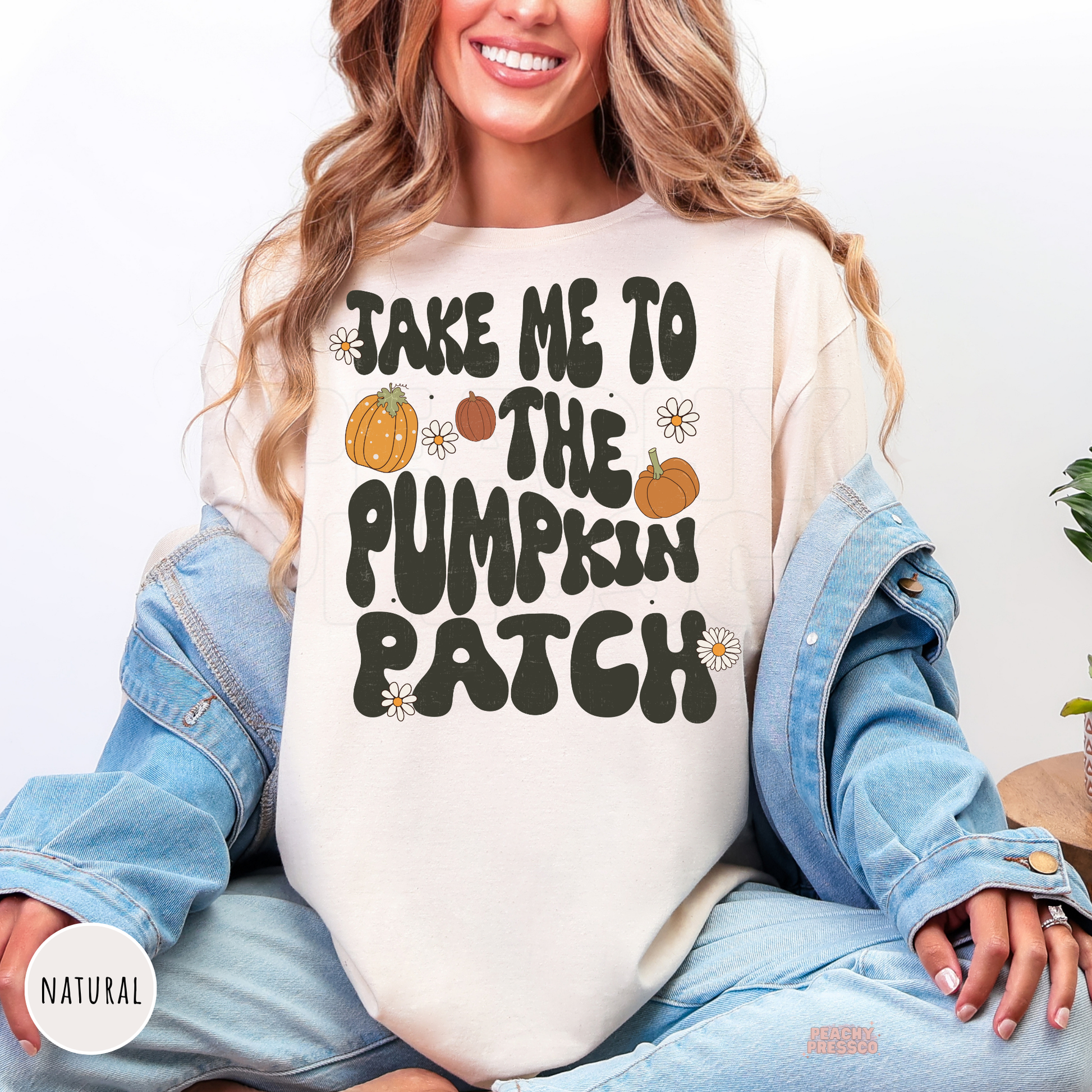 Woman wearing a natural graphic t-shirt featuring the phrase "Take Me to the Pumpkin Patch" with pumpkins and daisy flowers.