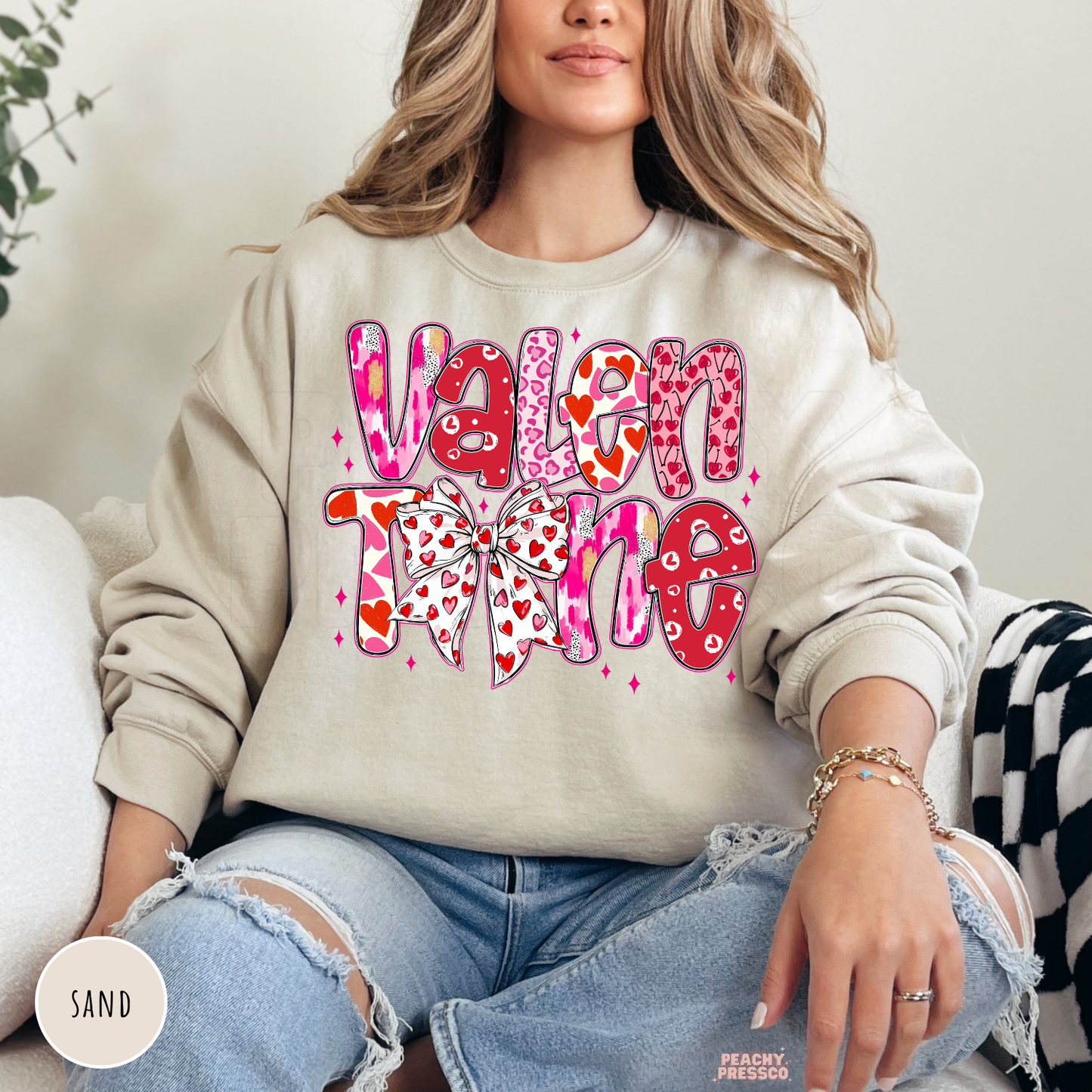 Sand crewneck sweatshirt displaying a stacked "Valentine" graphic with pink hearts and a decorative coquette bow.
