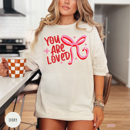 Ivory unisex Valentine's Day t-shirt with a vintage "You Are Loved" quote and pink ribbon bow graphic.