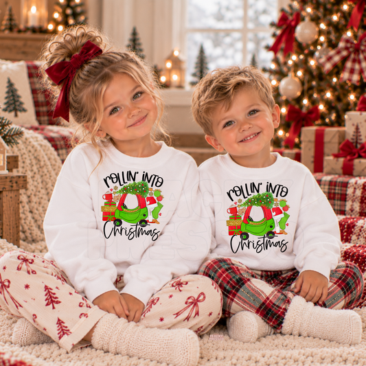 Boy and girl wearing white Rollin’ Into Christmas kids crewnecks with festive green car, presents and holiday design