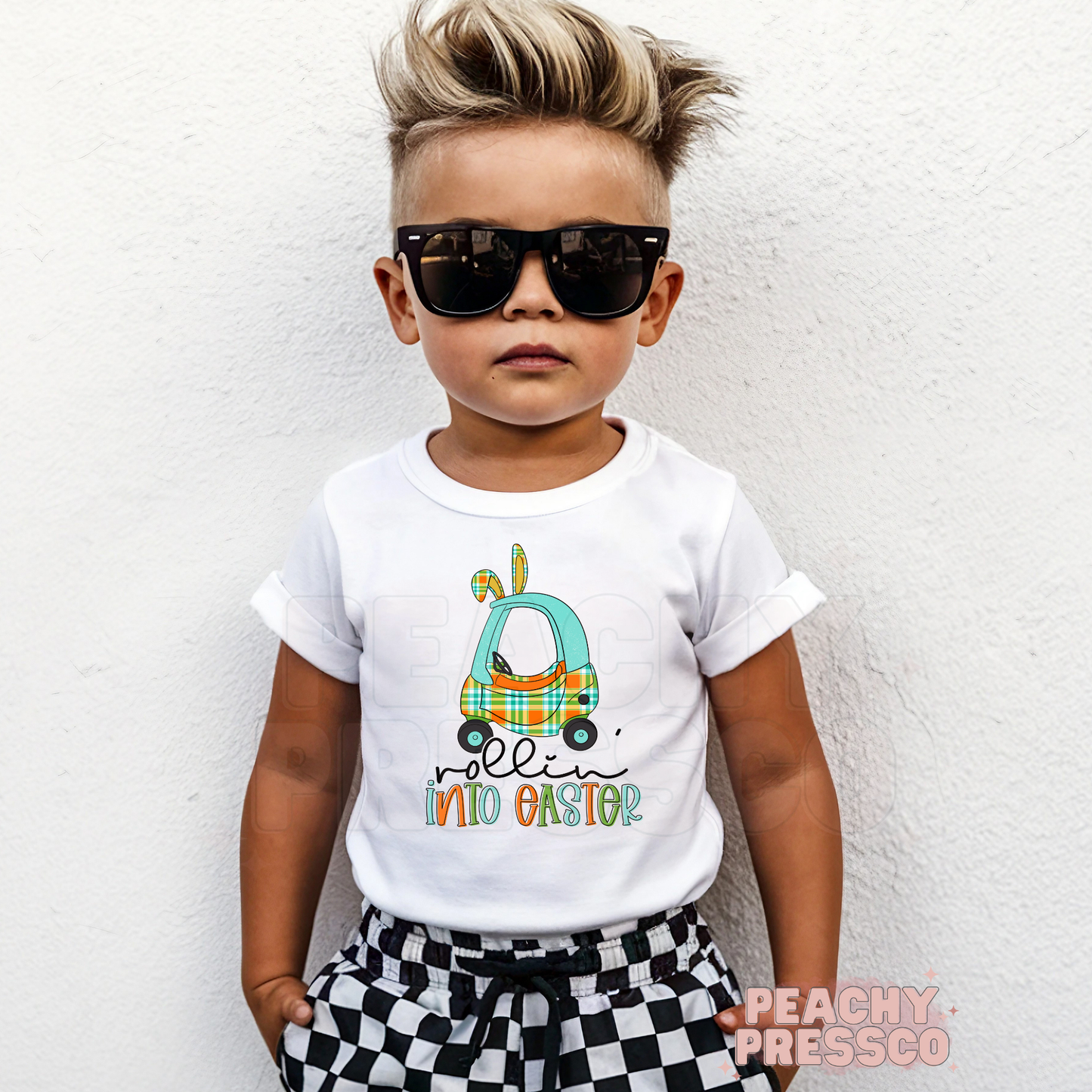 Toddler boy wearing a white Rollin Into Easter shirt featuring a blue plaid bunny car Easter graphic with bunny ears.