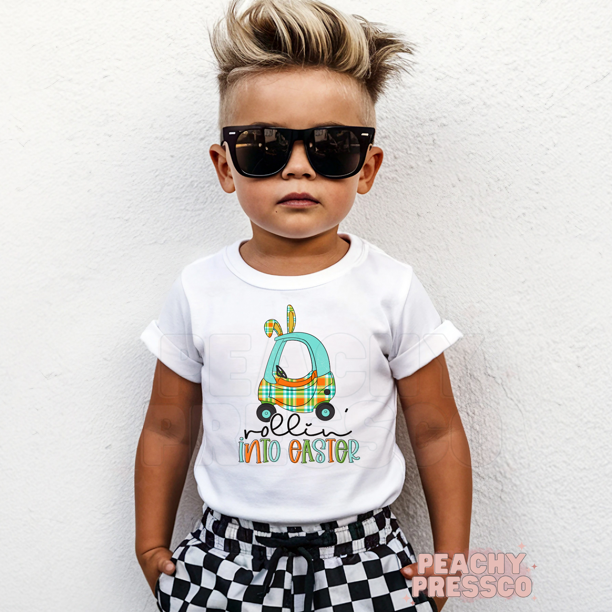 Toddler boy wearing a white Rollin Into Easter shirt featuring a blue plaid bunny car Easter graphic with bunny ears.