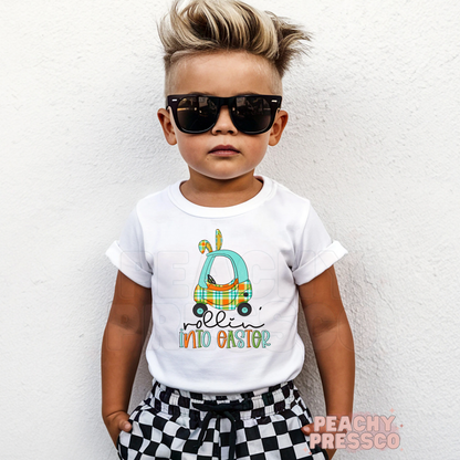 Toddler boy wearing a white Rollin Into Easter shirt featuring a blue plaid bunny car Easter graphic with bunny ears.