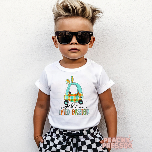 Toddler boy wearing a white Rollin Into Easter shirt featuring a blue plaid bunny car Easter graphic with bunny ears.